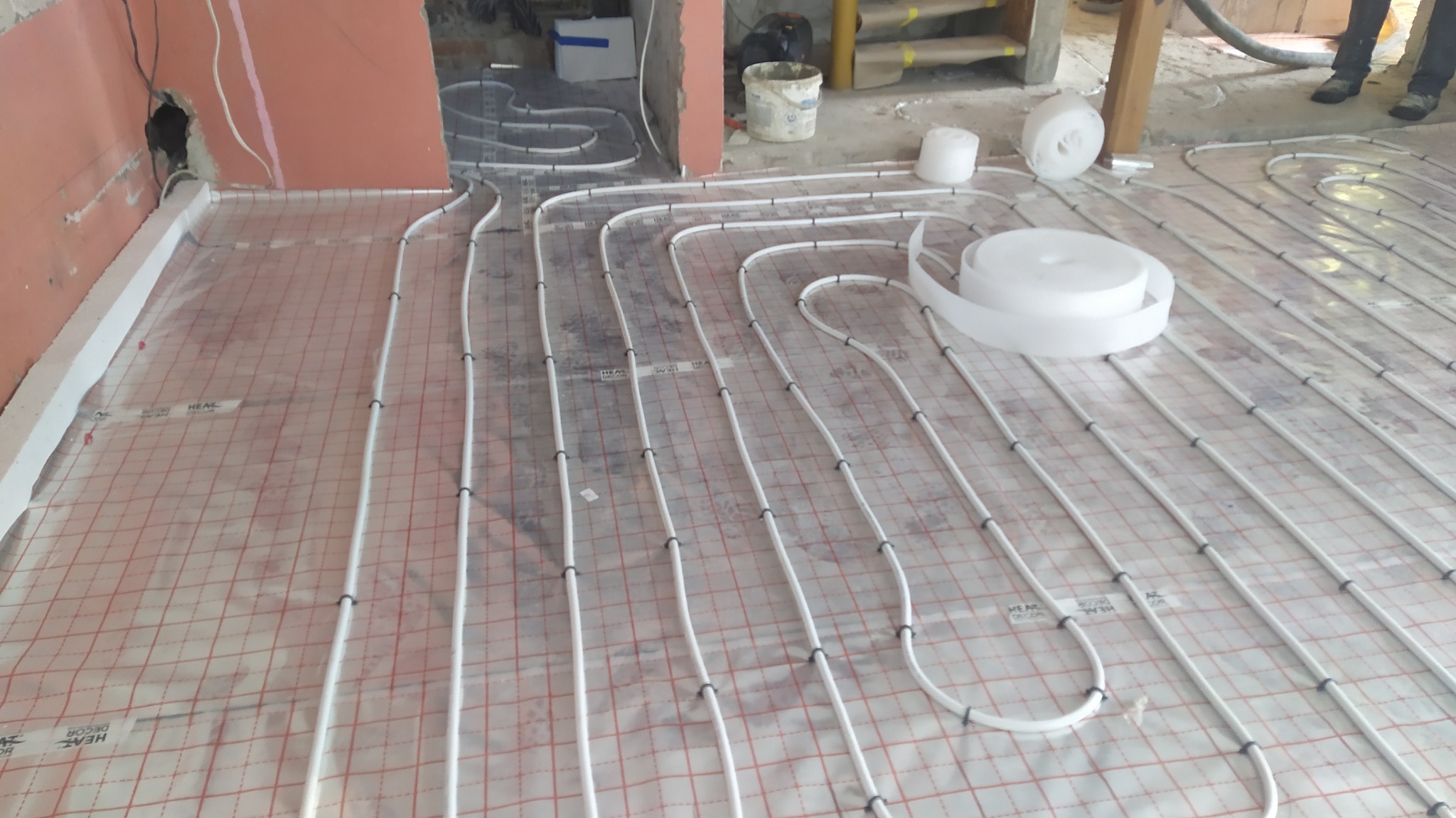 Rozłożona instalacja ogrzewania podłogowego na folii z siatką, widoczne rury mocowane spinkami, fragment ściany w stanie surowym oraz rolka taśmy montażowej.