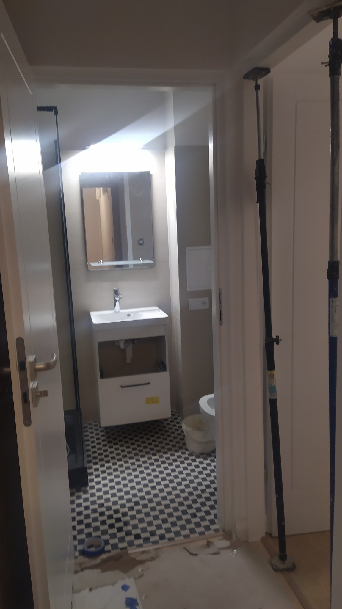 Łazienka w trakcie remontu: widok na umywalkę z szafką i lustrem, czarno-białą podłogę w szachownicę, oraz wsporniki budowlane podtrzymujące ościeżnicę drzwiową.