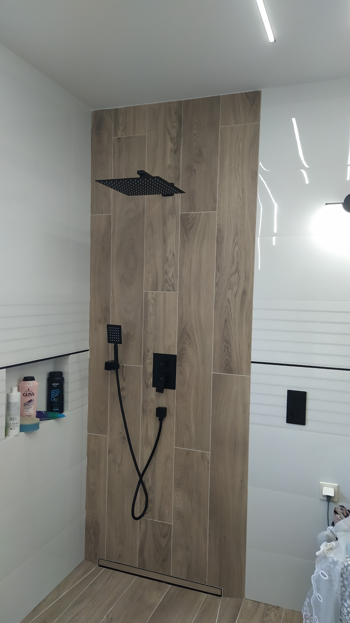Nowoczesna kabina prysznicowa z czarną armaturą, drewnopodobnymi płytkami na ścianie i odpływem liniowym w podłodze, uzupełniona o półkę z kosmetykami.