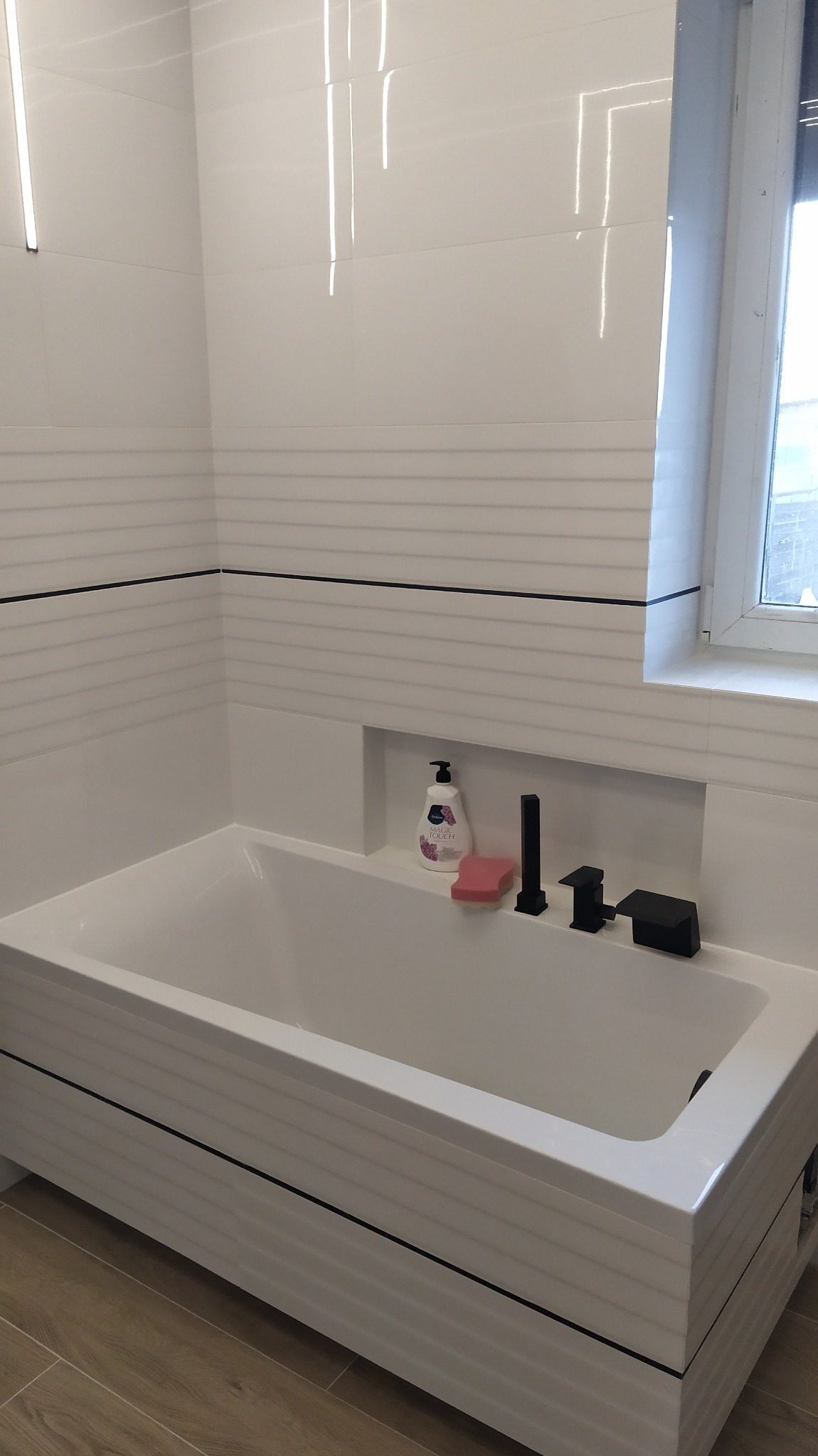 Nowoczesna, biała wanna z czarną armaturą i półką z kosmetykami w łazience wykończonej białymi płytkami z poziomymi liniami.