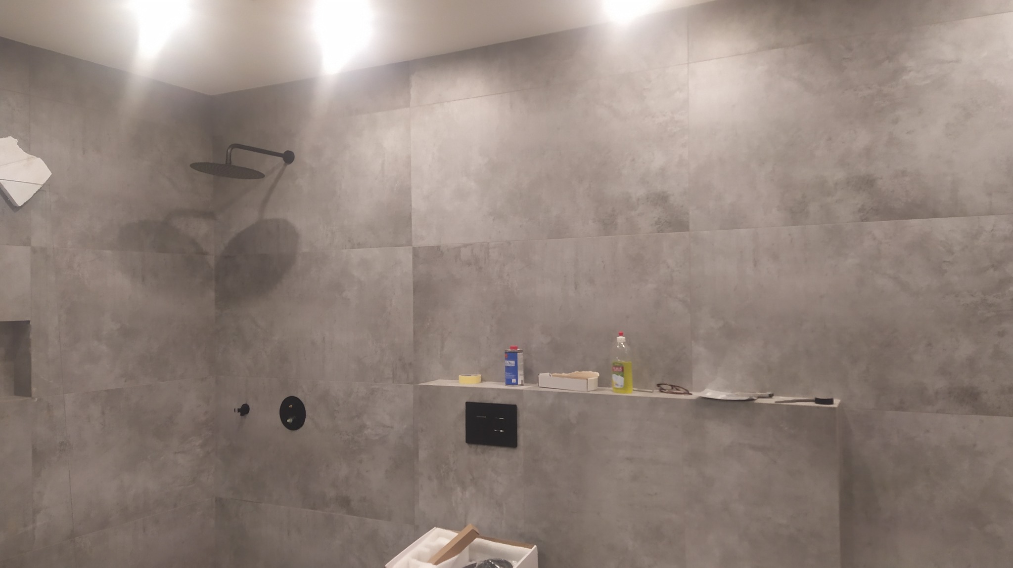 Wykończona łazienka z szarymi płytkami imitującymi beton, czarną armaturą i półką z akcesoriami oraz materiałami budowlanymi w trakcie prac wykończeniowych.