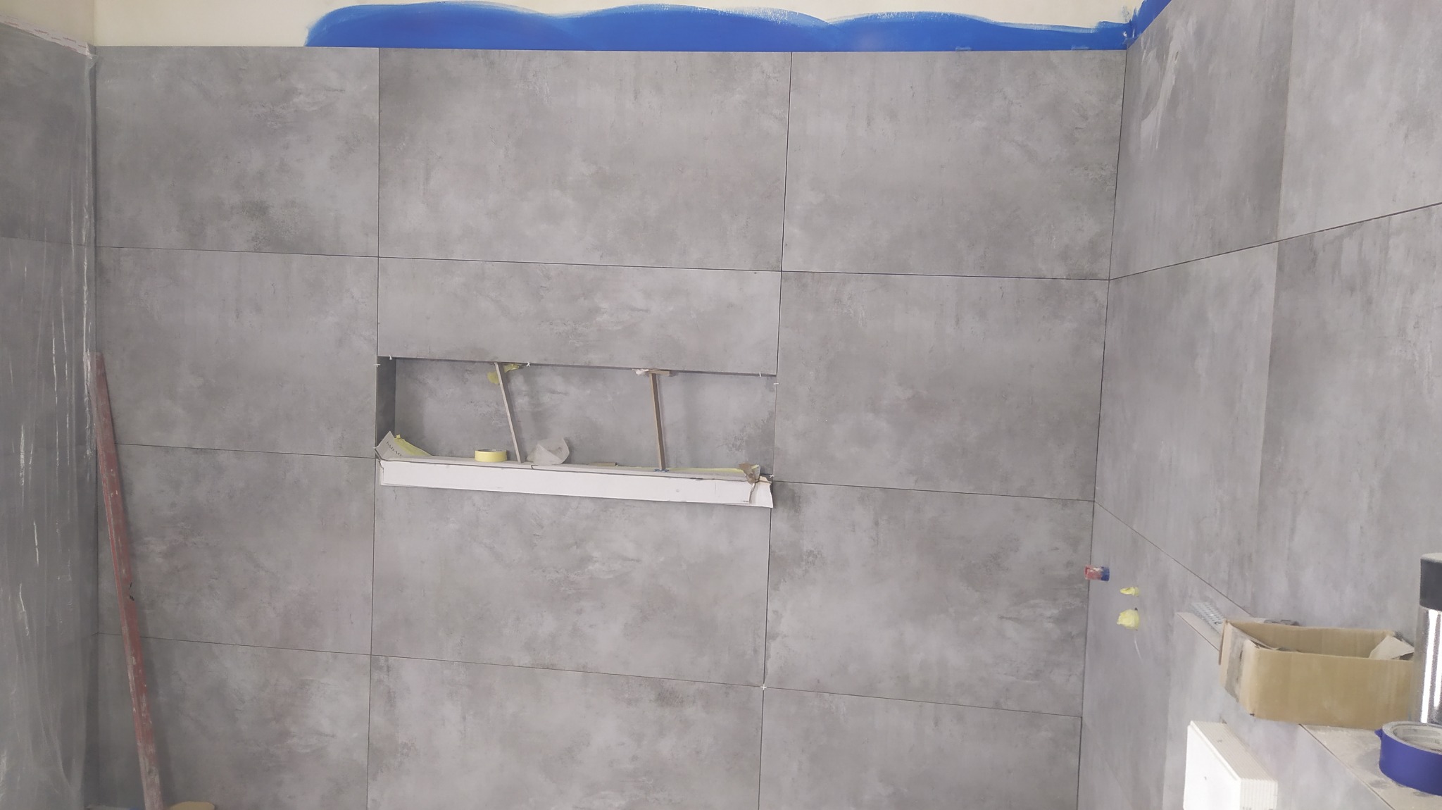 Ściana łazienki wyłożona szarymi płytkami w trakcie montażu, widoczna wnęka z prowizorycznym wsparciem, zabezpieczona folią i taśmą malarską