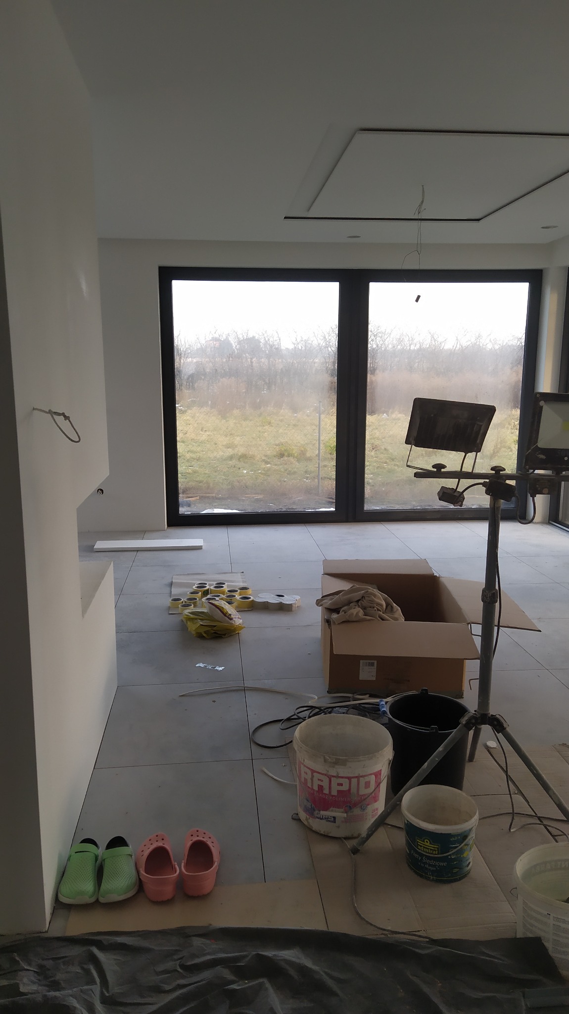 Wnętrze w trakcie wykańczania: widoczne gładkie, białe ściany, duże okno z czarną ramą, szare płytki na podłodze, rozłożone narzędzia i materiały budowlane, w tym puszki z farbą, taśmy malarskie...