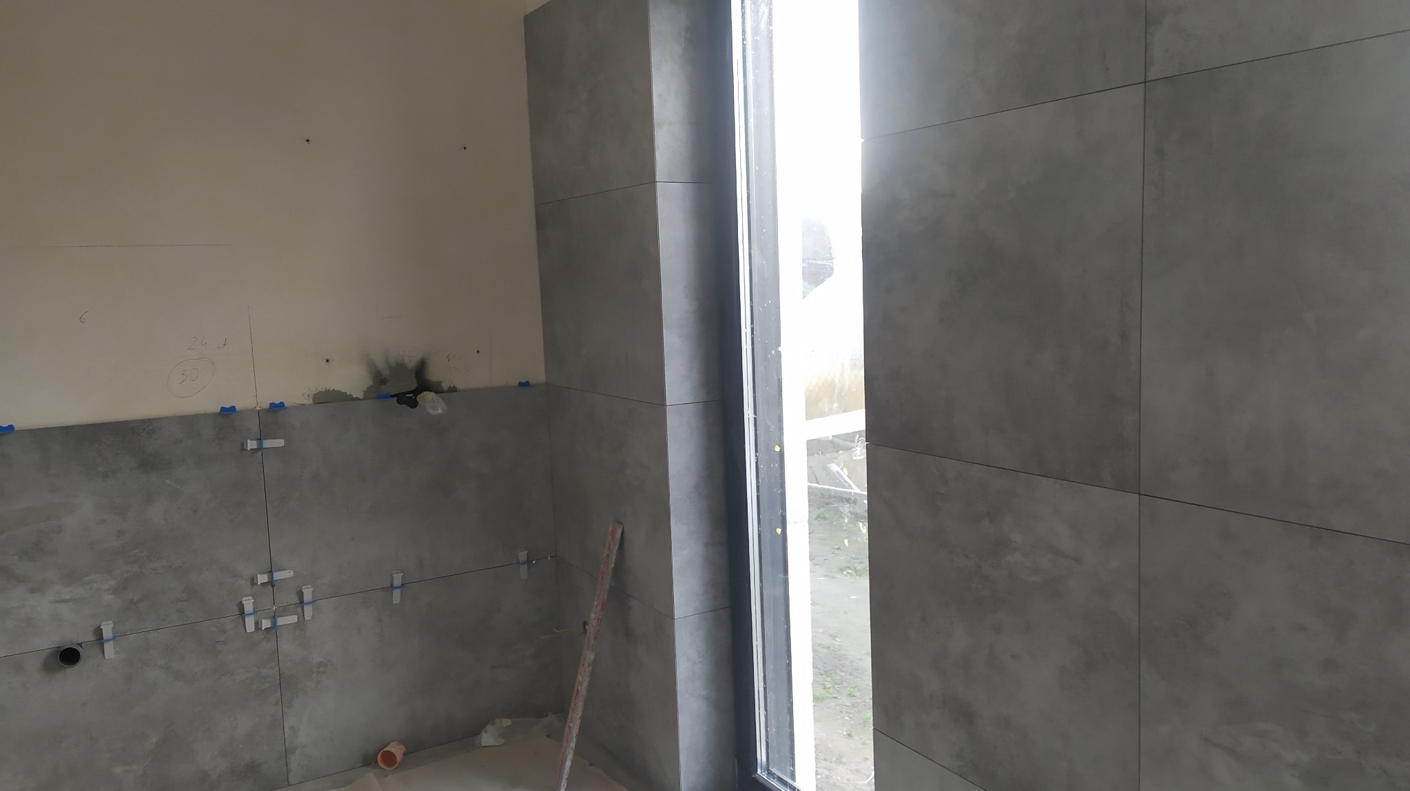 Szare płytki ścienne w trakcie montażu, widoczne plastikowe krzyżyki dystansowe, pionowa linia na ścianie z numerem 30, okno z widokiem na zewnątrz.