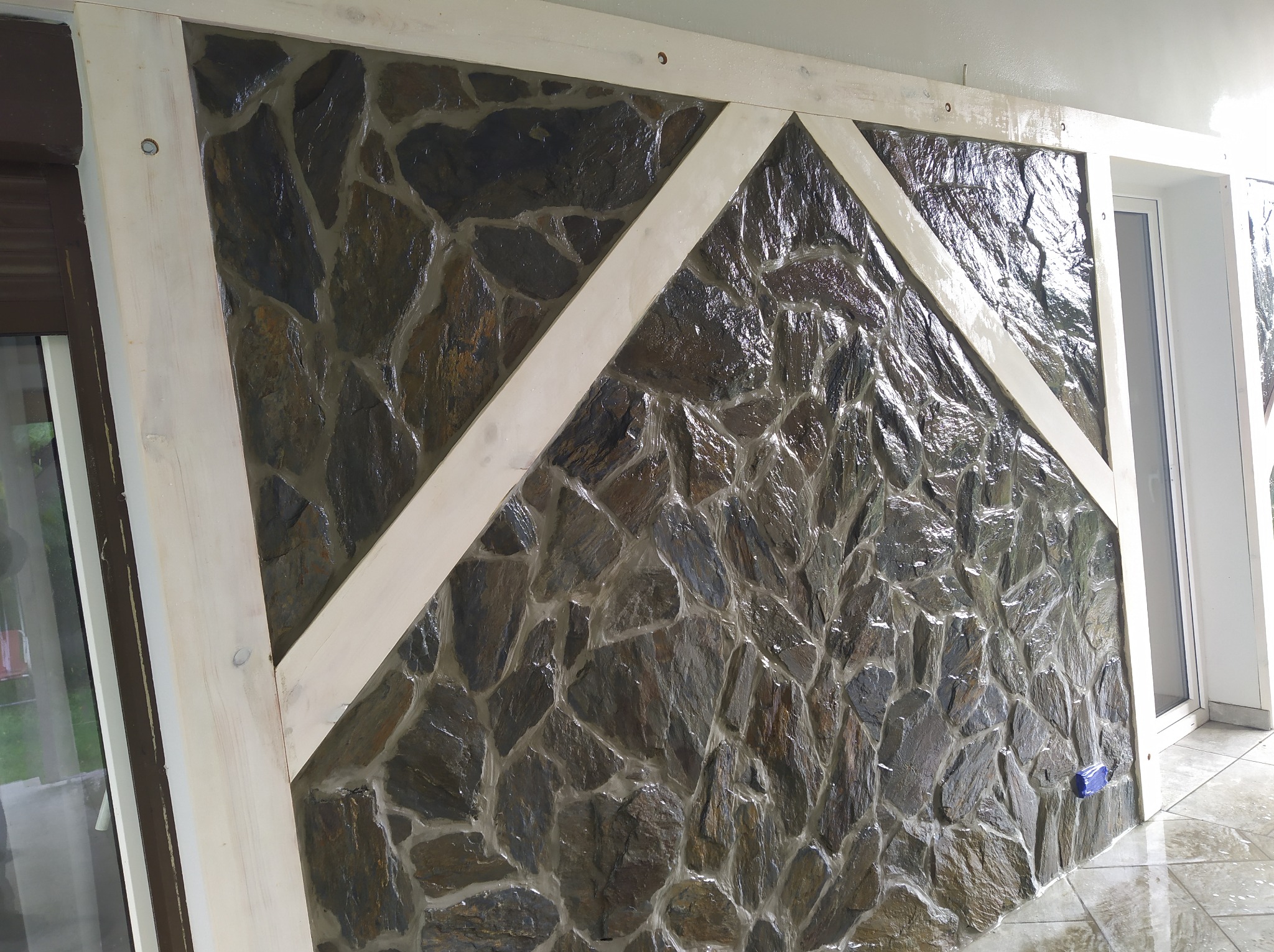 Ściana wyłożona nieregularnym kamieniem w odcieniach szarości i brązu, z drewnianą ramą w jasnym odcieniu, tworzącą geometryczny wzór na powierzchni