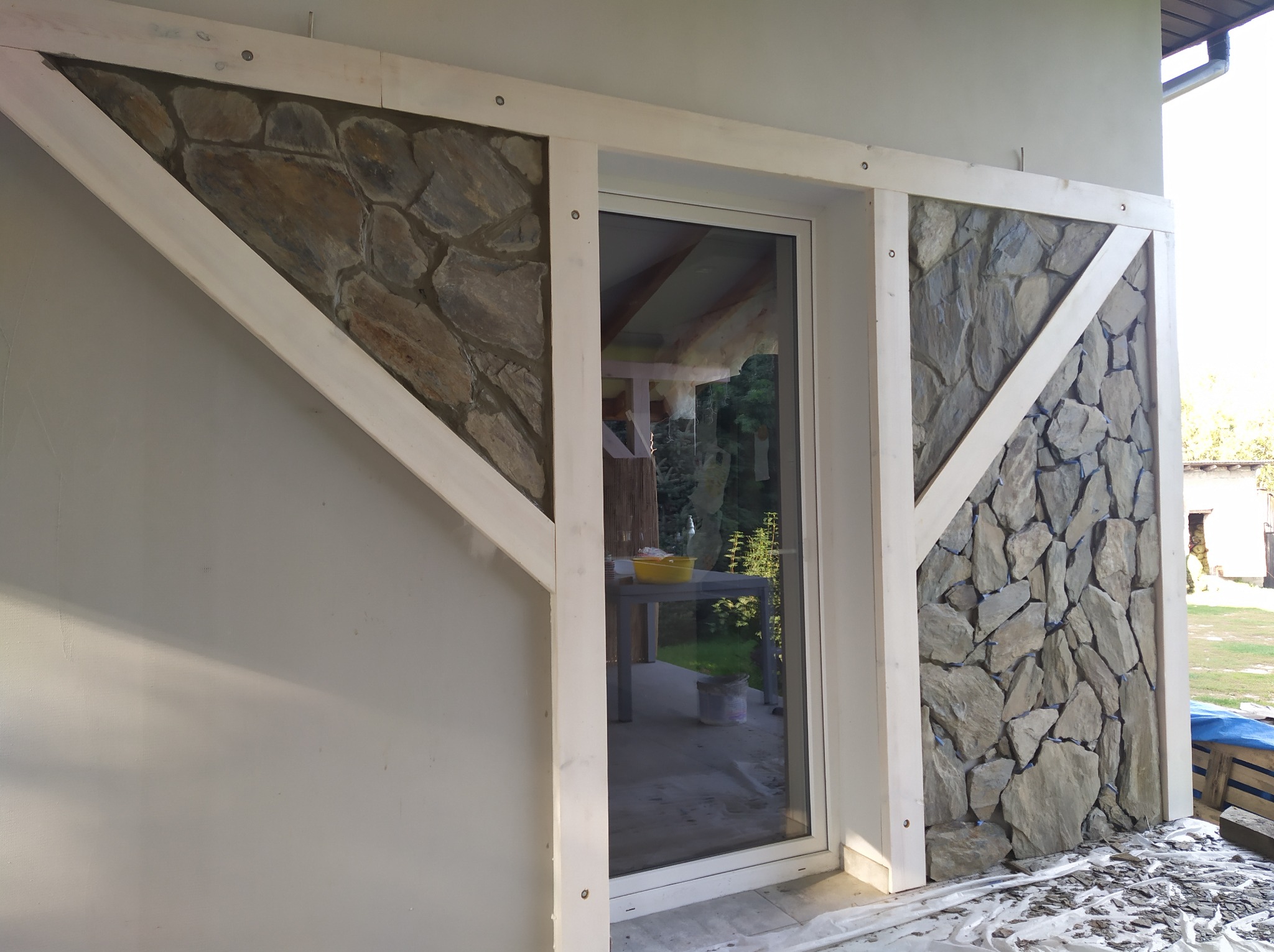 Fragment elewacji budynku z wykończeniem z kamienia naturalnego w drewnianej ramie, widoczny przez okno stół z żółtą miską i wiadro, na podłożu folia ochronna.