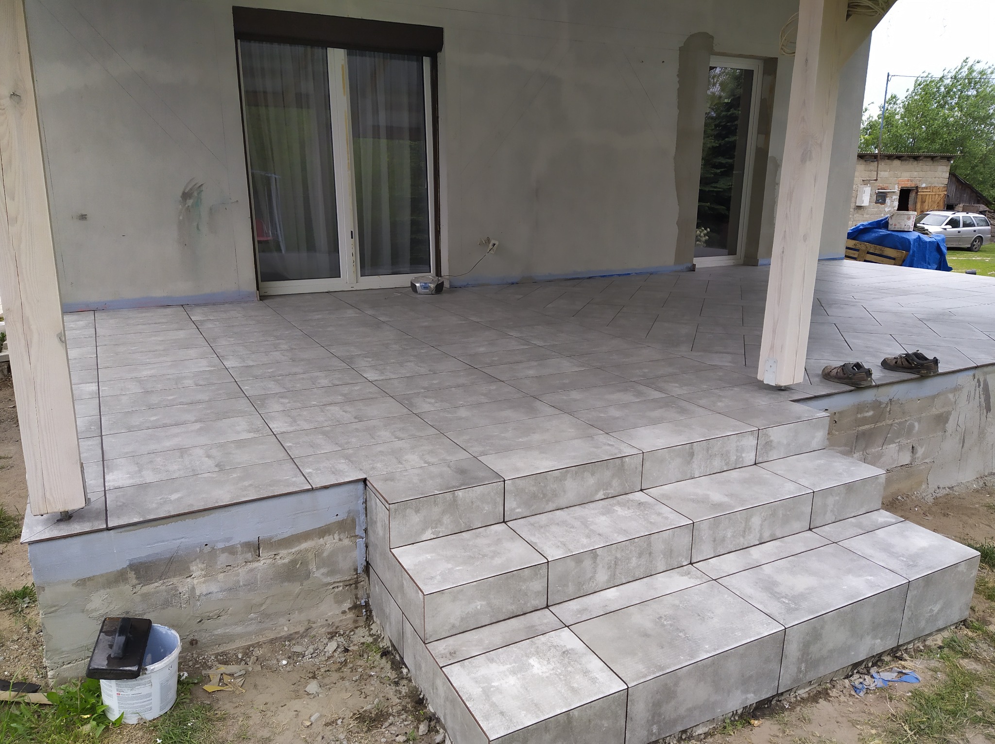 Wykończony taras z szarymi płytkami i schodami, widoczny biały słup podtrzymujący zadaszenie, na podłodze leżą buty robocze, wiadro i narzędzia budowlane.
