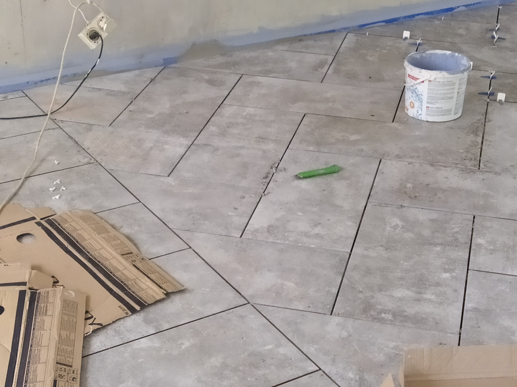 Układanie szarych płytek podłogowych w jodełkę, widoczny fragment ściany z niebieską taśmą malarską, wiadro po farbie i kartony po płytkach.