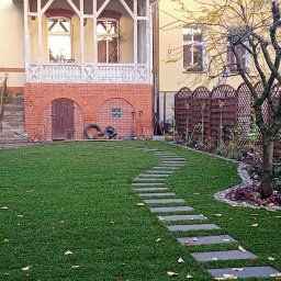 Jan Jaślanek - Ułożona trawa z rolki w ogrodzie z drewnianym ogrodzeniem i ścieżką z płyt. W tle budynek z ceglanymi elementami i balkonem. Jesienne liście na trawie.