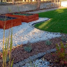 Jan Jaślanek - Ogród z podwyższonymi rabatami, białym żwirem i obrzeżami z kostki. Widoczna trawa, kora i rośliny ozdobne. Ujęcie z perspektywy ziemi, podkreślające detale.