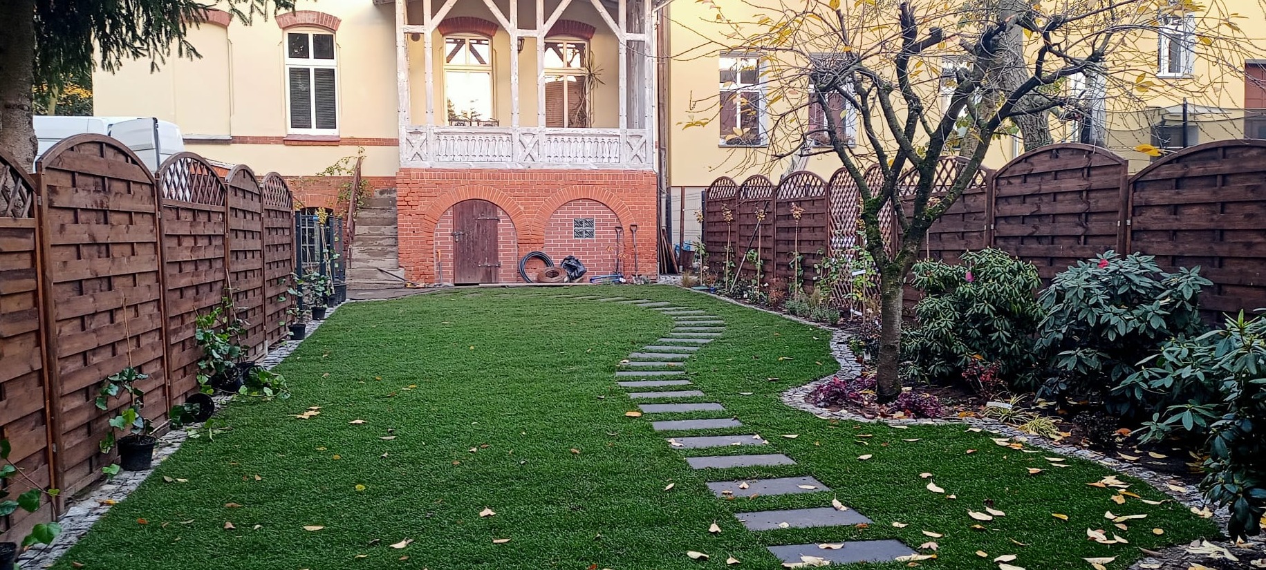 Ułożona trawa z rolki w ogrodzie z drewnianym ogrodzeniem i ścieżką z płyt. W tle budynek z ceglanymi elementami i balkonem. Jesienne liście na trawie.