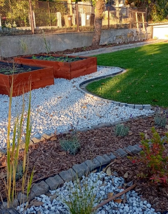 Ogród z podwyższonymi rabatami, białym żwirem i obrzeżami z kostki. Widoczna trawa, kora i rośliny ozdobne. Ujęcie z perspektywy ziemi, podkreślające detale.