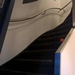 Rob-Kop - Ciemne schody z metalową poręczą i dwoma abstrakcyjnymi obrazami na ścianie. Ujęcie z dołu, widoczne stopnie i fragment ściany.