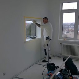 Rob-Kop - Malarz w białym kombinezonie maluje ramę okienną w jasnym pokoju, zabezpieczoną taśmą malarską. Widoczne narzędzia i wiadro z farbą. Za oknem zabudowa miejska.