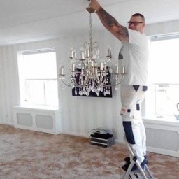 Rob-Kop - Mężczyzna na drabinie demontuje kryształowy żyrandol w pokoju z białymi ścianami i brązowym dywanem. Widoczne okno i grzejnik pod nim.