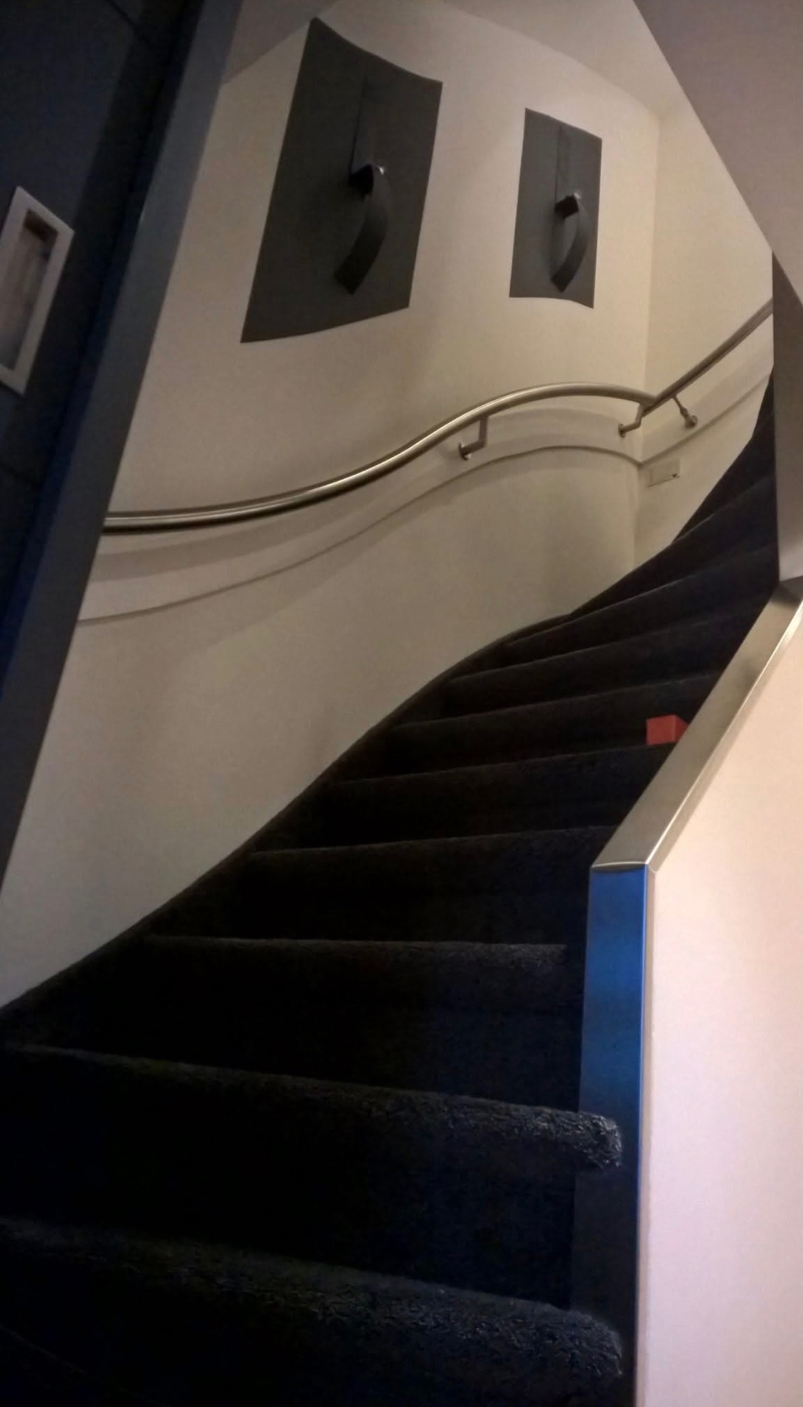 Ciemne schody z metalową poręczą i dwoma abstrakcyjnymi obrazami na ścianie. Ujęcie z dołu, widoczne stopnie i fragment ściany.