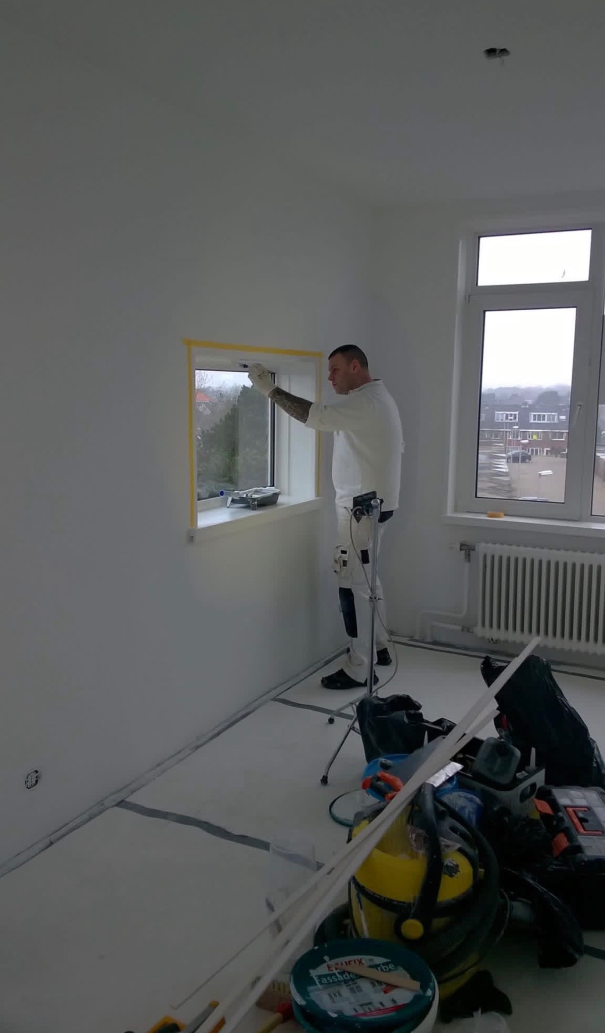 Malarz w białym kombinezonie maluje ramę okienną w jasnym pokoju, zabezpieczoną taśmą malarską. Widoczne narzędzia i wiadro z farbą. Za oknem zabudowa miejska.