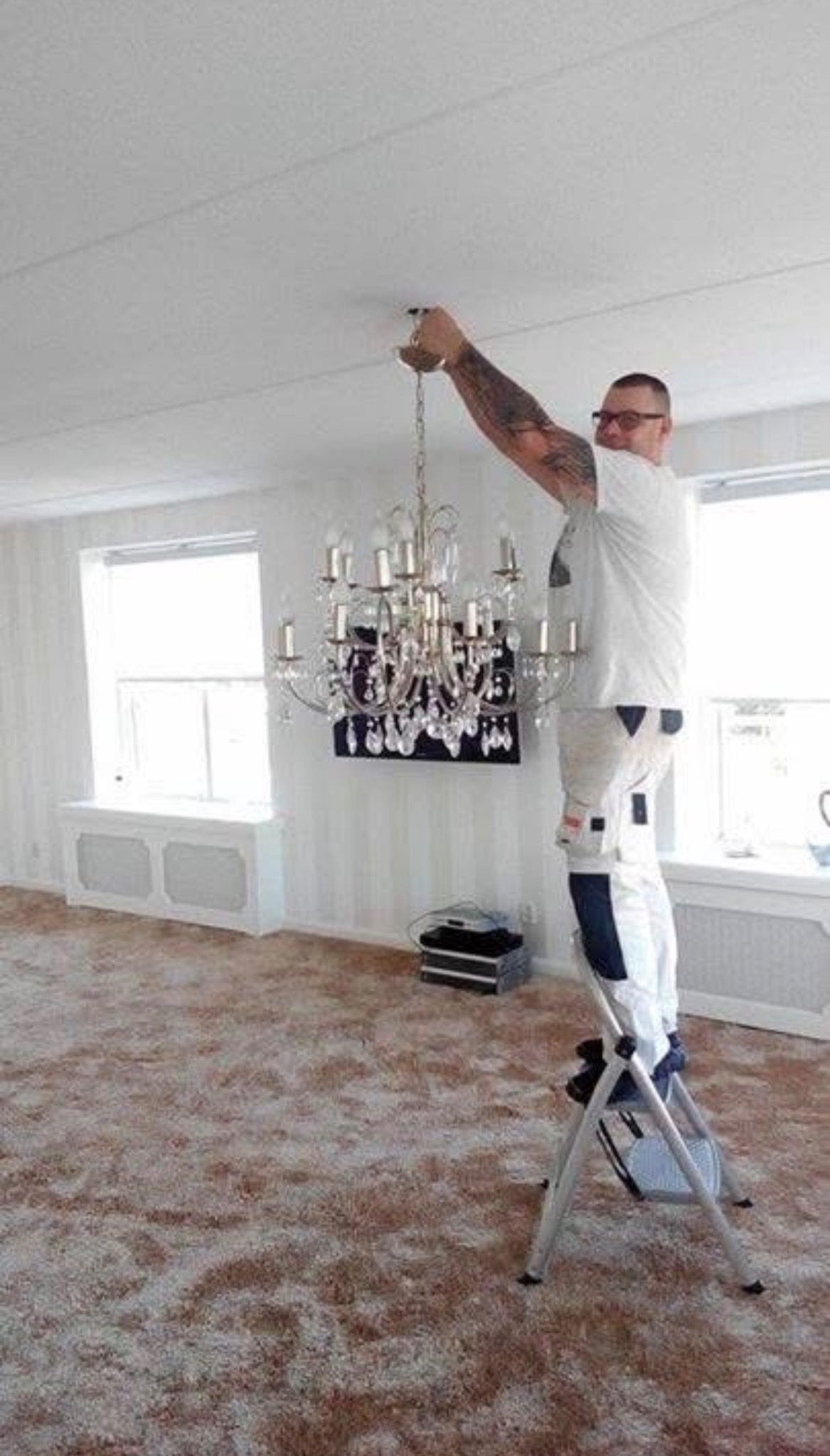 Mężczyzna na drabinie demontuje kryształowy żyrandol w pokoju z białymi ścianami i brązowym dywanem. Widoczne okno i grzejnik pod nim.