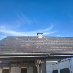 Klaudia Rusiecka - Nowy, grafitowy dach z dachówki ceramicznej, z zamontowanymi rynnami, na tle błękitnego nieba. Widoczny komin i fragment budynku w trakcie budowy.