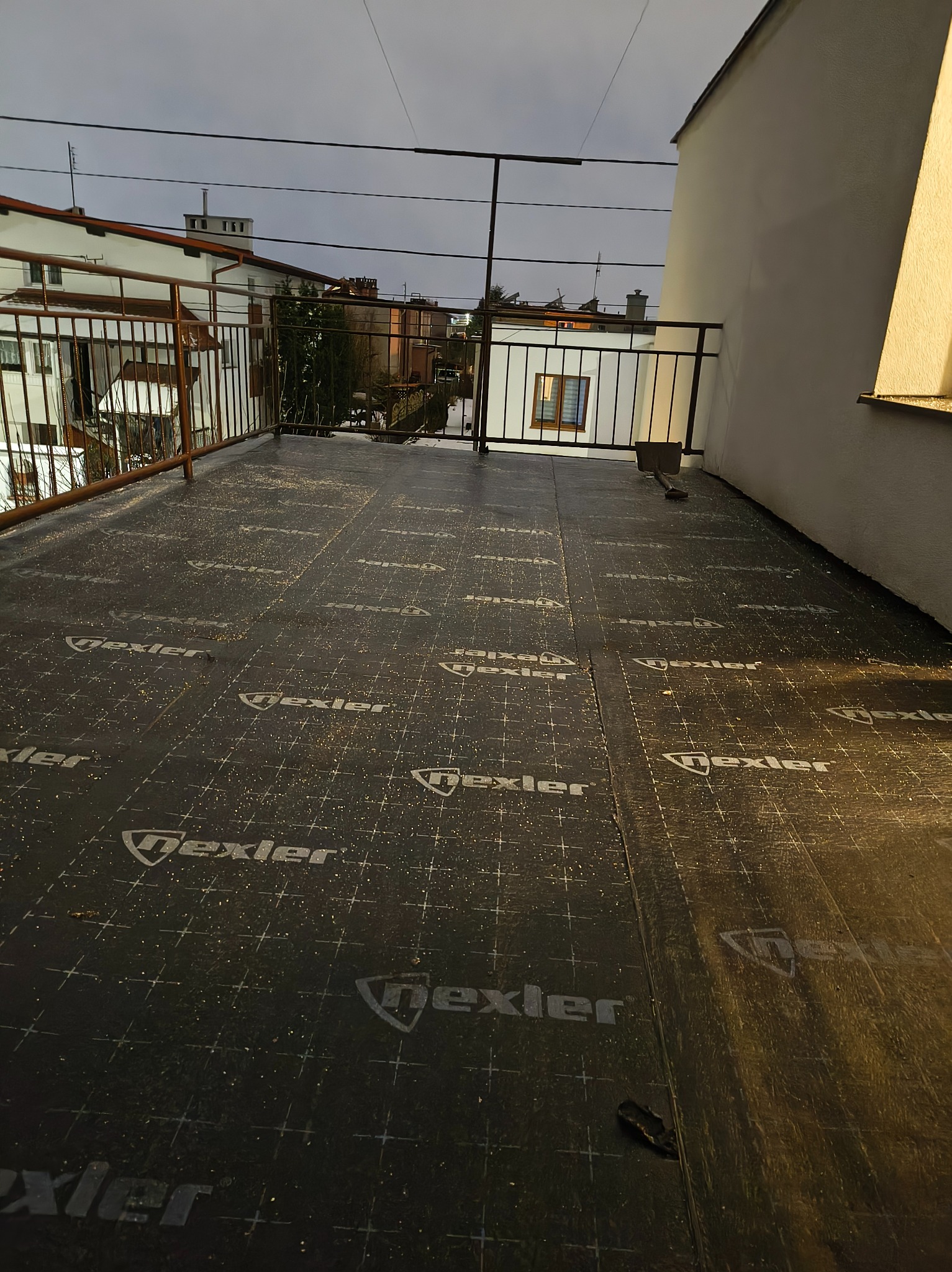 Wykończony dach płaski z czarną membraną hydroizolacyjną marki Dexler, zabezpieczony balustradą. Widok z góry na tle zabudowy miejskiej w pochmurny dzień.