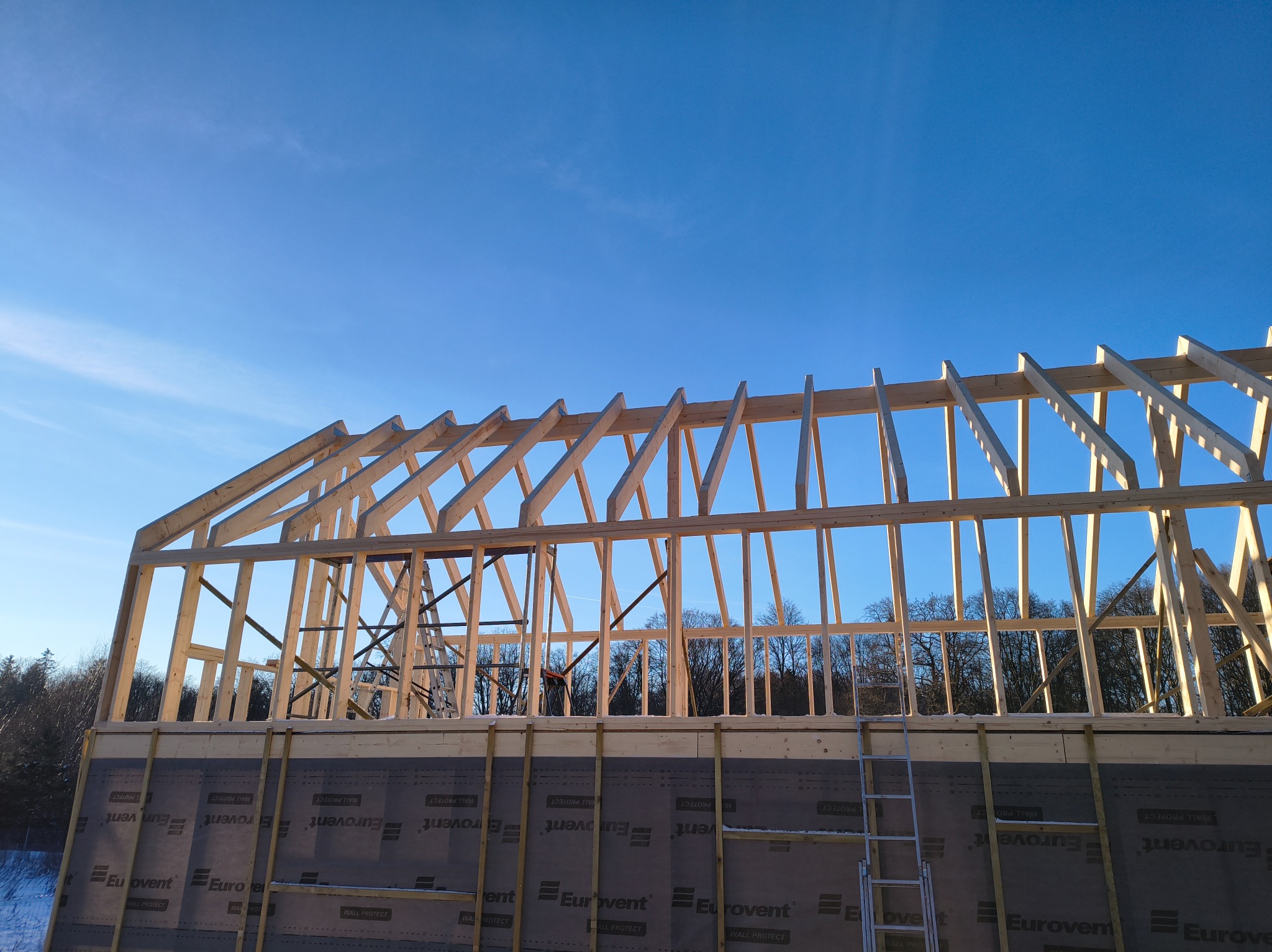 Konstrukcja drewniana dachu w trakcie budowy, widok z dołu na tle błękitnego nieba. Widoczna drabina oparta o ścianę z membraną Eurovent.