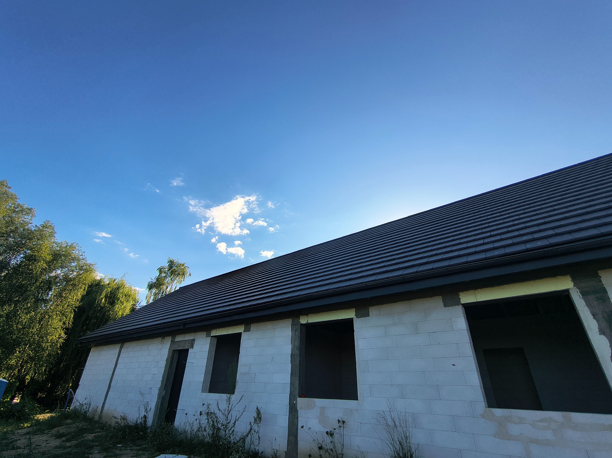 Nowy, ciemny dach z rynnami na surowym budynku z białego bloczka, błękitne niebo w tle. Widok z dołu, zieleń drzew po lewej stronie.