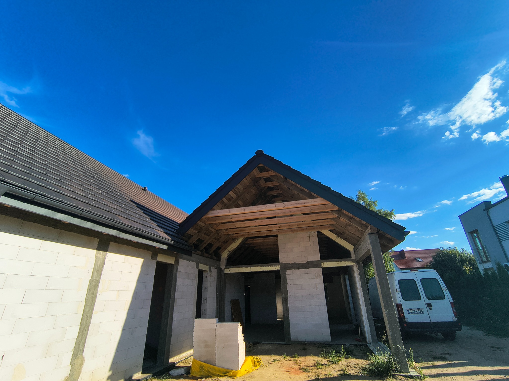Budowa domu jednorodzinnego w trakcie realizacji. Widoczny fragment dachu z czarną dachówką, ściany z białego bloczka i drewniana konstrukcja zadaszenia.