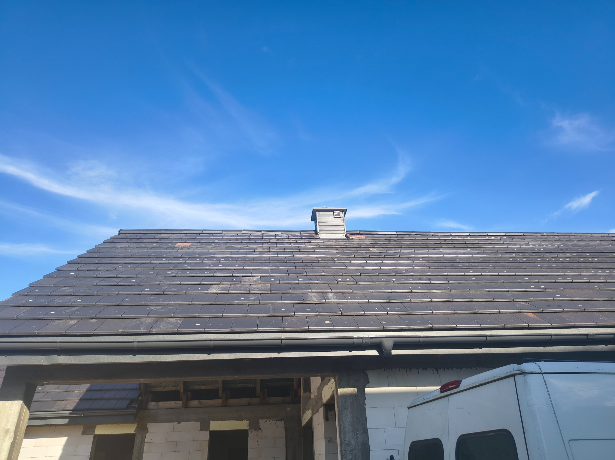 Nowy, grafitowy dach z dachówki ceramicznej, z zamontowanymi rynnami, na tle błękitnego nieba. Widoczny komin i fragment budynku w trakcie budowy.