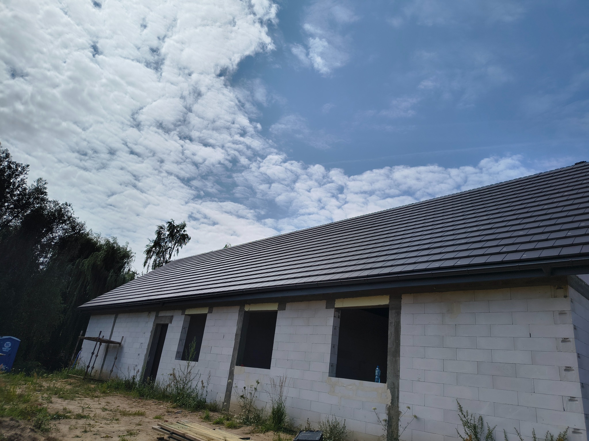 Nowy, ciemny dach z dachówki ceramicznej na surowym budynku z betonu komórkowego w słoneczny dzień. Widoczne niezabudowane otwory okienne i fragment lasu w tle.