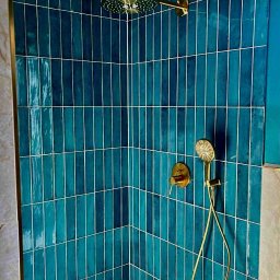 Andrzej Połata - Kabina prysznicowa z turkusowymi, pionowo ułożonymi płytkami i złotą armaturą. Widoczny odpływ liniowy i fragment marmurowej ściany.
