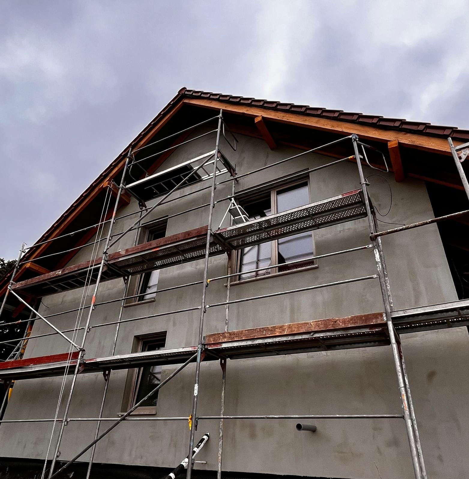 Dom jednorodzinny w trakcie prac elewacyjnych, z widocznymi rusztowaniami i szarą, surową elewacją. Widok z dołu na budynek w pochmurny dzień.