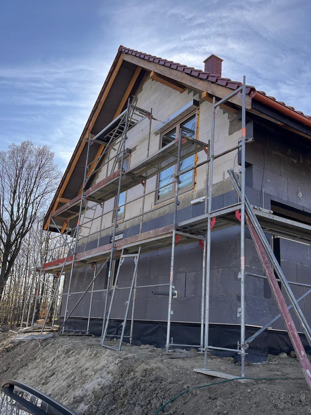 Dom w trakcie ocieplania, widoczne rusztowania, styropian grafitowy i białe bloczki. Częściowo ocieplona elewacja, okna z ramami. W tle drzewa i błękitne niebo.