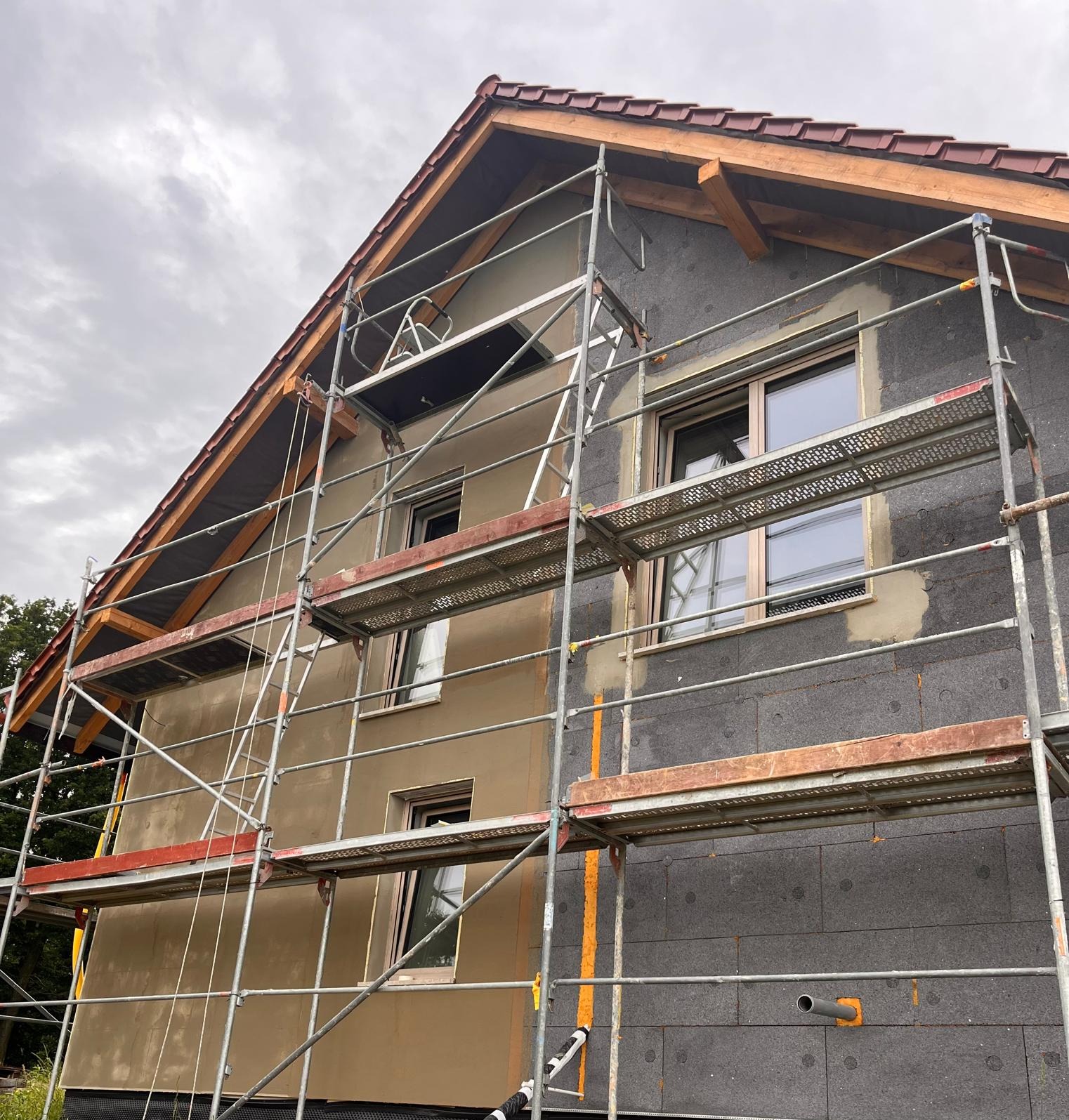 Dom w trakcie ocieplania: część ściany z szarym styropianem, część z nałożonym brązowym tynkiem. Widoczne rusztowania i okna. Pochmurne niebo w tle.