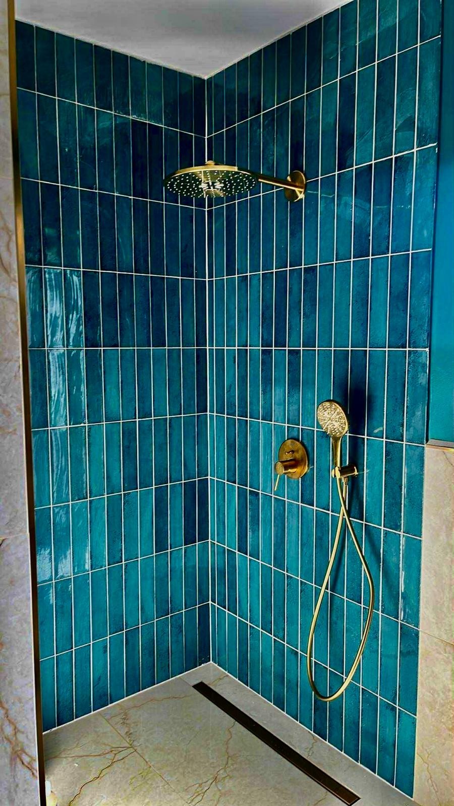 Kabina prysznicowa z turkusowymi, pionowo ułożonymi płytkami i złotą armaturą. Widoczny odpływ liniowy i fragment marmurowej ściany.
