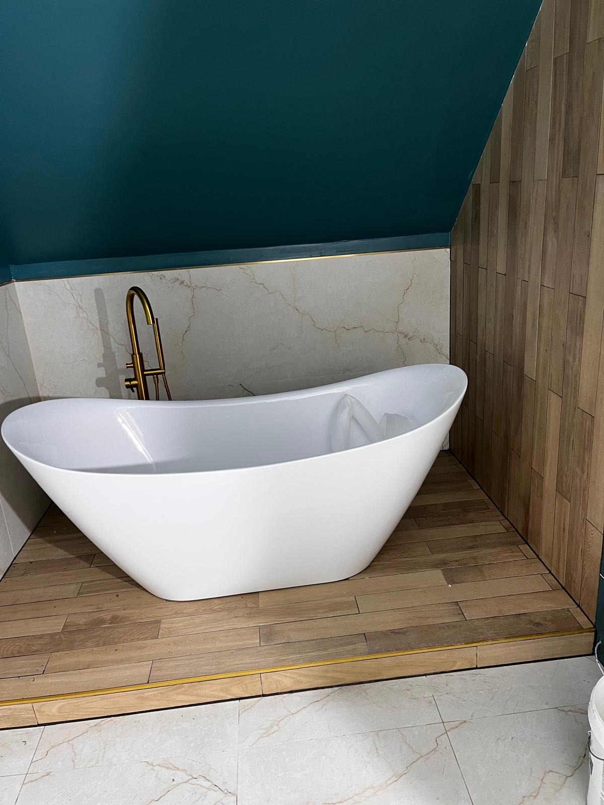 Nowoczesna, wolnostojąca wanna w łazience na poddaszu z drewnianą okładziną i złotą armaturą. Ściany wykończone płytkami imitującymi marmur.