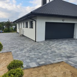 Naimad - Nowoczesny podjazd z szarej kostki brukowej ułożonej we wzór, z widocznym fragmentem przygotowanego podłoża i elementami zieleni. Dom z garażem w tle.