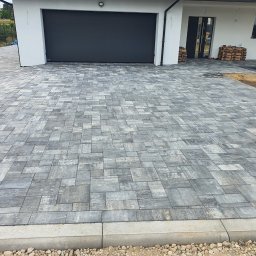 Naimad - Szara kostka brukowa ułożona na podjeździe przed nowoczesnym domem z garażem. Widoczne worki z materiałami budowlanymi i obrzeża z kamykami.