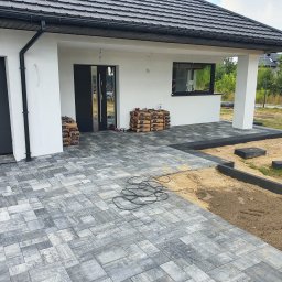 Naimad - Nowoczesny podjazd z szarej kostki brukowej przed domem. Częściowo ułożona nawierzchnia, widoczne worki z materiałem i kable. Obrzeża z kostki. Etap prac.