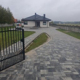 Naimad - Posesja z nowoczesnym domem i podjazdem z szarej kostki brukowej, widok zza otwartej, czarnej bramy. W tle ogrodzenie i zaparkowany samochód dostawczy.