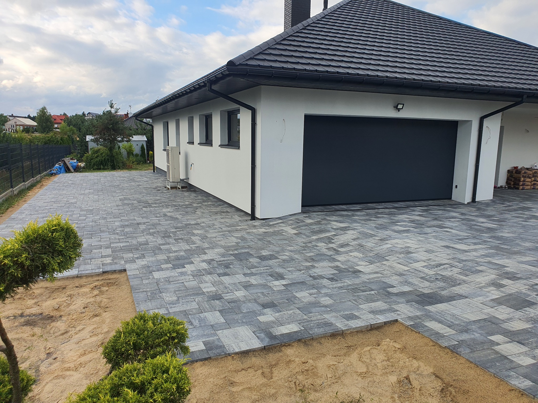 Nowoczesny podjazd z szarej kostki brukowej ułożonej we wzór, z widocznym fragmentem przygotowanego podłoża i elementami zieleni. Dom z garażem w tle.