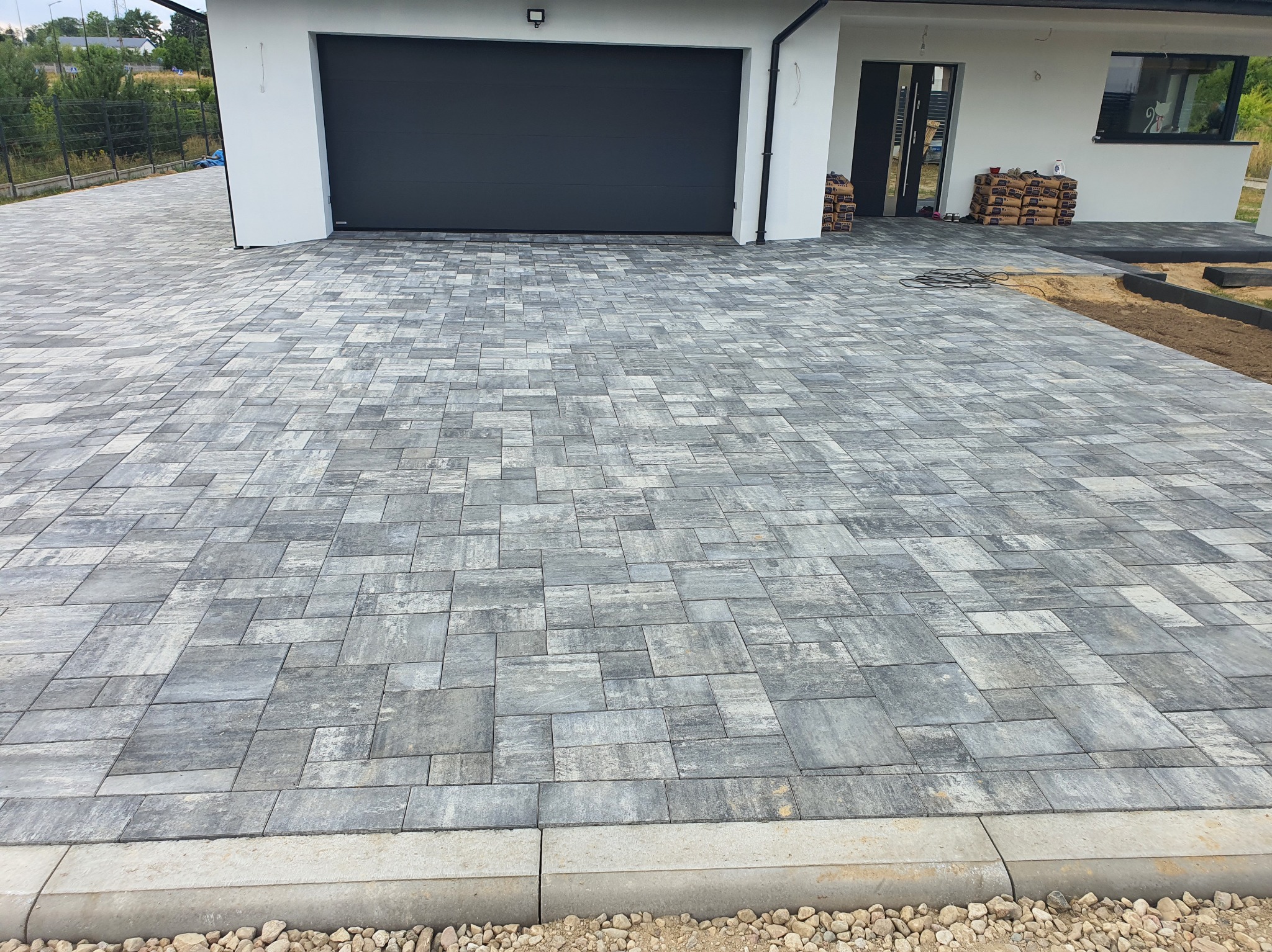 Szara kostka brukowa ułożona na podjeździe przed nowoczesnym domem z garażem. Widoczne worki z materiałami budowlanymi i obrzeża z kamykami.