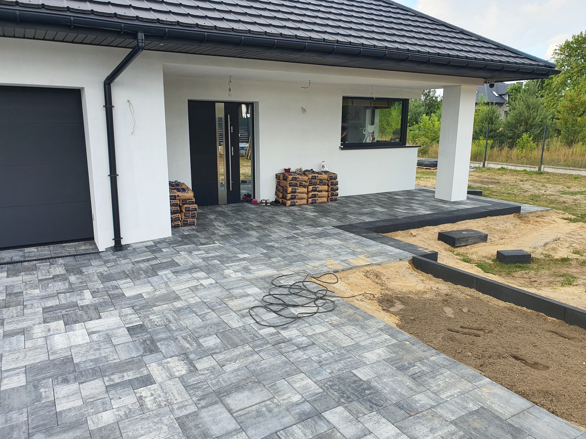 Nowoczesny podjazd z szarej kostki brukowej przed domem. Częściowo ułożona nawierzchnia, widoczne worki z materiałem i kable. Obrzeża z kostki. Etap prac.
