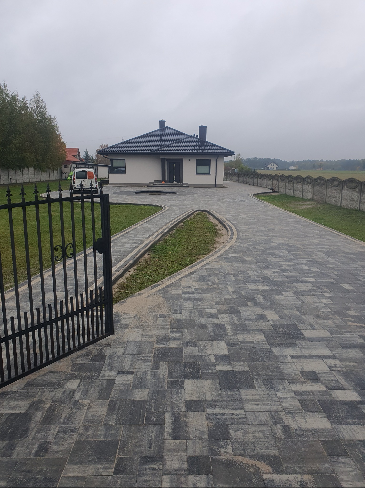 Posesja z nowoczesnym domem i podjazdem z szarej kostki brukowej, widok zza otwartej, czarnej bramy. W tle ogrodzenie i zaparkowany samochód dostawczy.