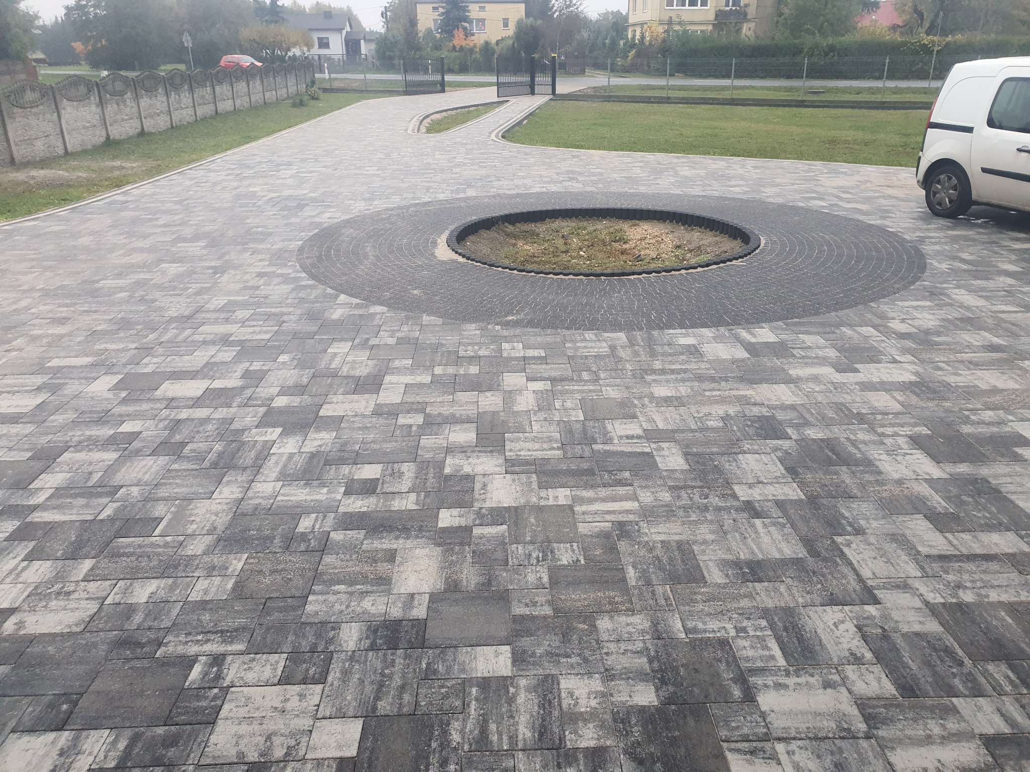 Nowoczesna nawierzchnia z kostki brukowej w odcieniach szarości, z okrągłym elementem centralnym i ścieżką prowadzącą do bramy. Widoczny fragment ogrodzenia i zaparkowany samochód.