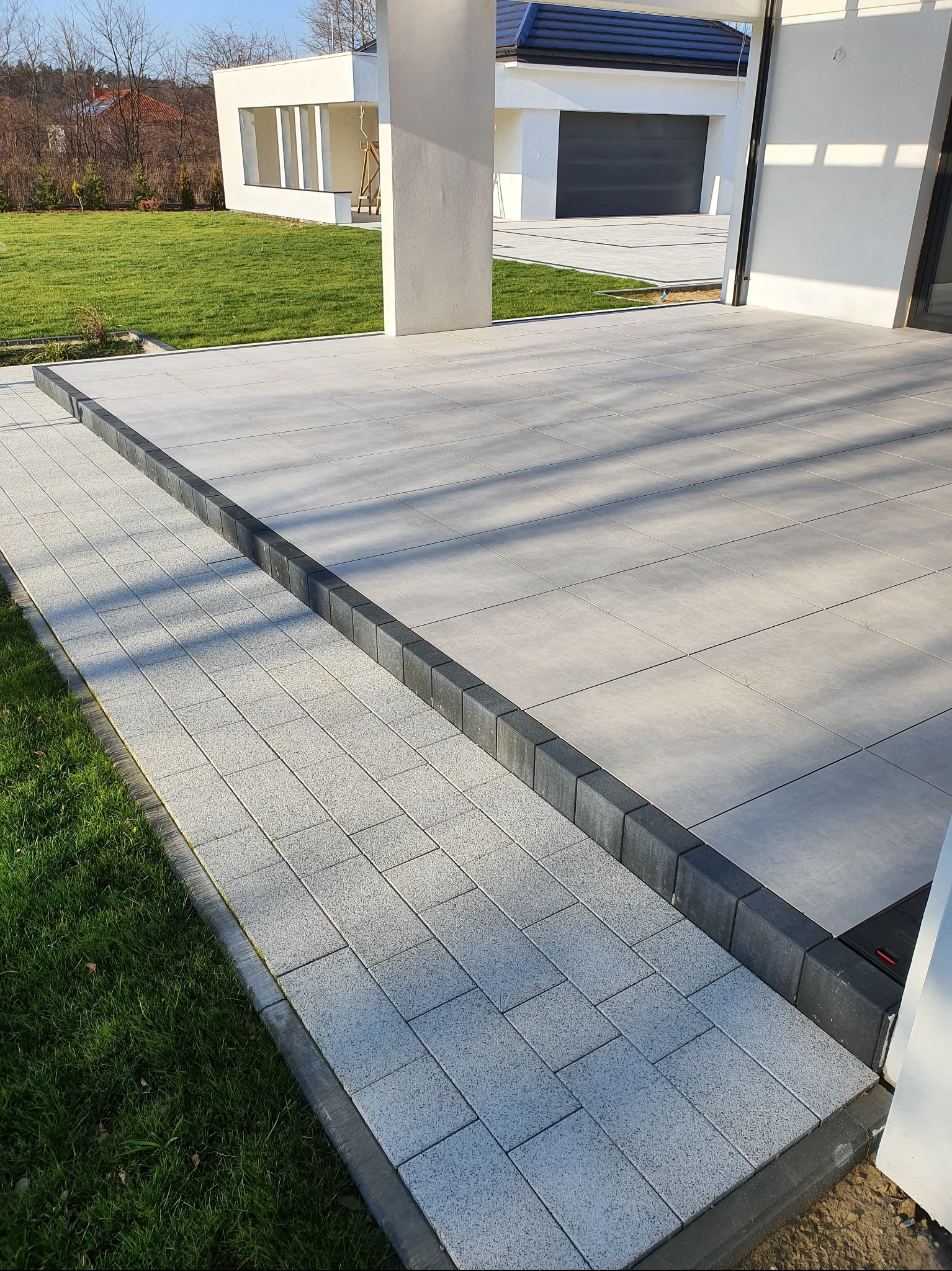 Nowoczesny taras z jasnoszarej kostki brukowej, obrzeża z ciemnoszarego betonu. W tle budynek z garażem i trawnik. Minimalistyczny design.