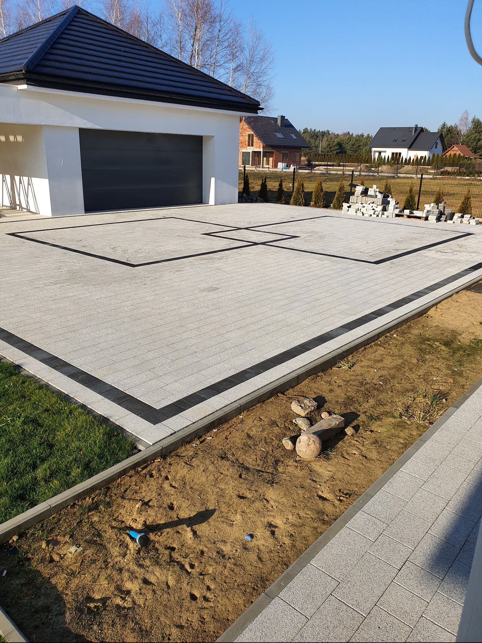 Jasnoszara kostka brukowa z czarnymi obramowaniami tworząca wzór na podjeździe. W tle dom z garażem i ułożone palety z kostką.