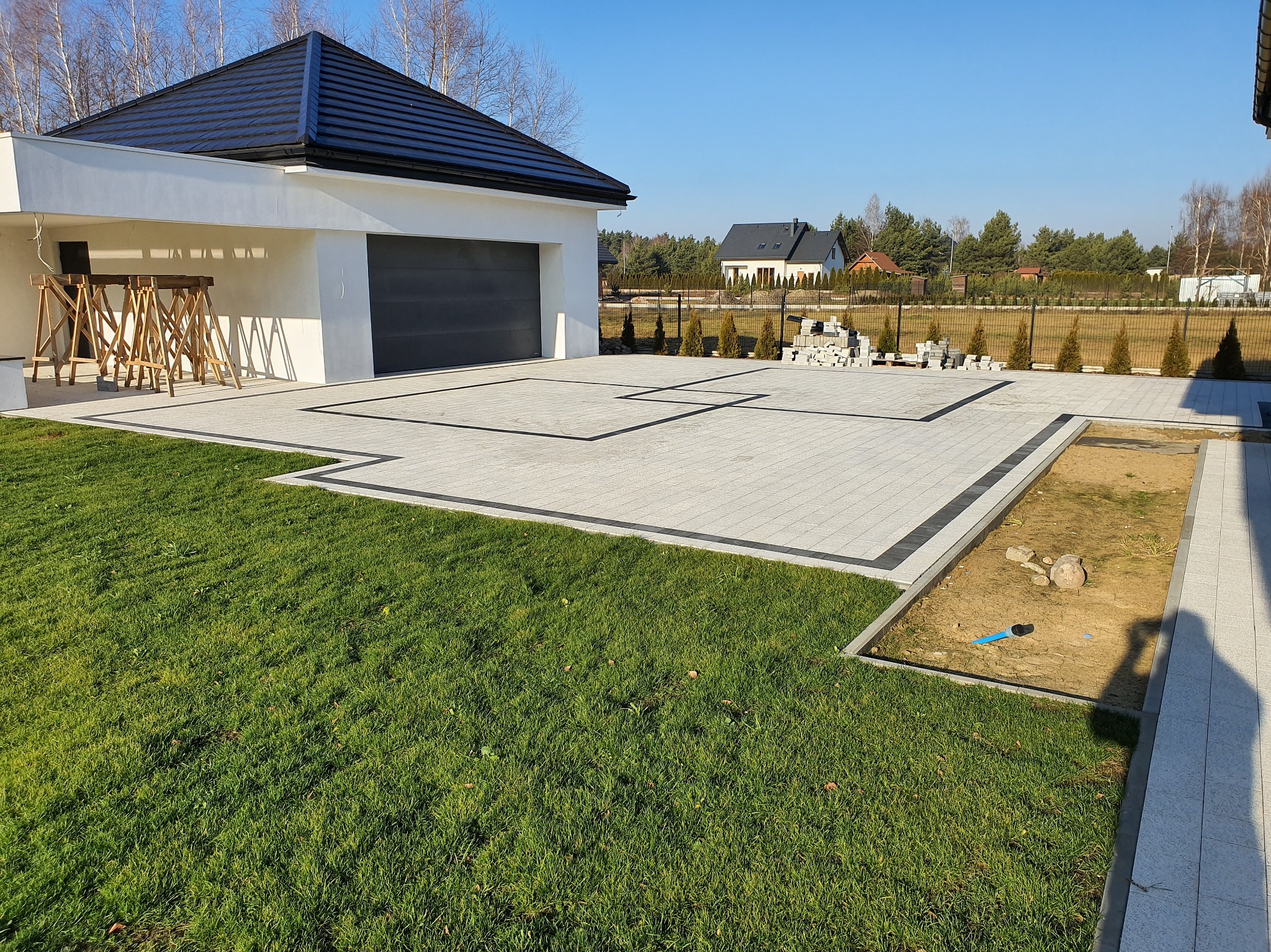 Jasnoszara kostka brukowa z czarną obwódką tworzy geometryczny wzór na podjeździe nowoczesnego domu z garażem. Część kostki jeszcze nieułożona. Trawa i ogrodzenie w tle.