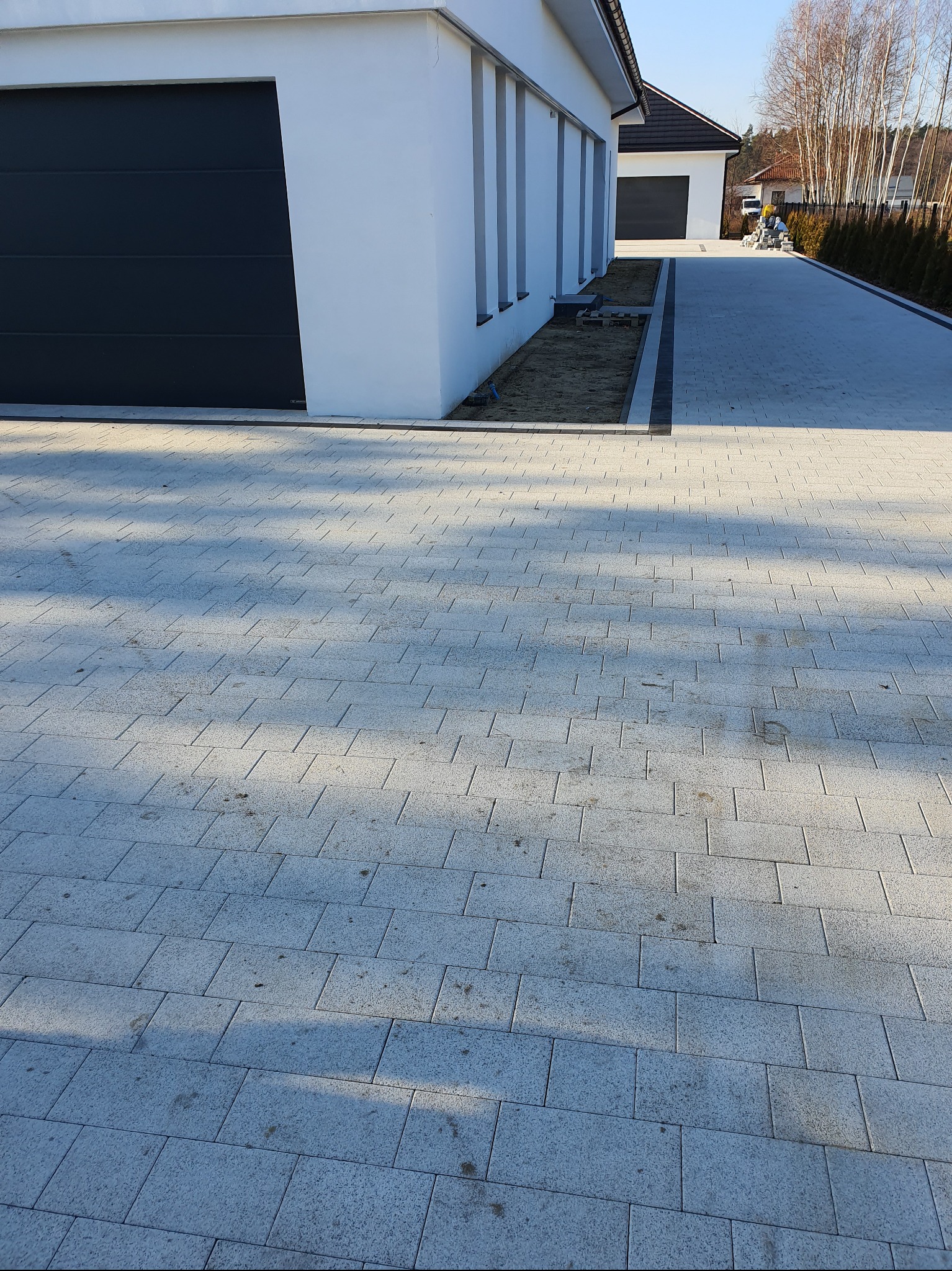 Jasnoszara kostka brukowa ułożona na podjeździe, widok z perspektywy, zarys budynku z garażem w tle, ciemna obwódka z kostki.