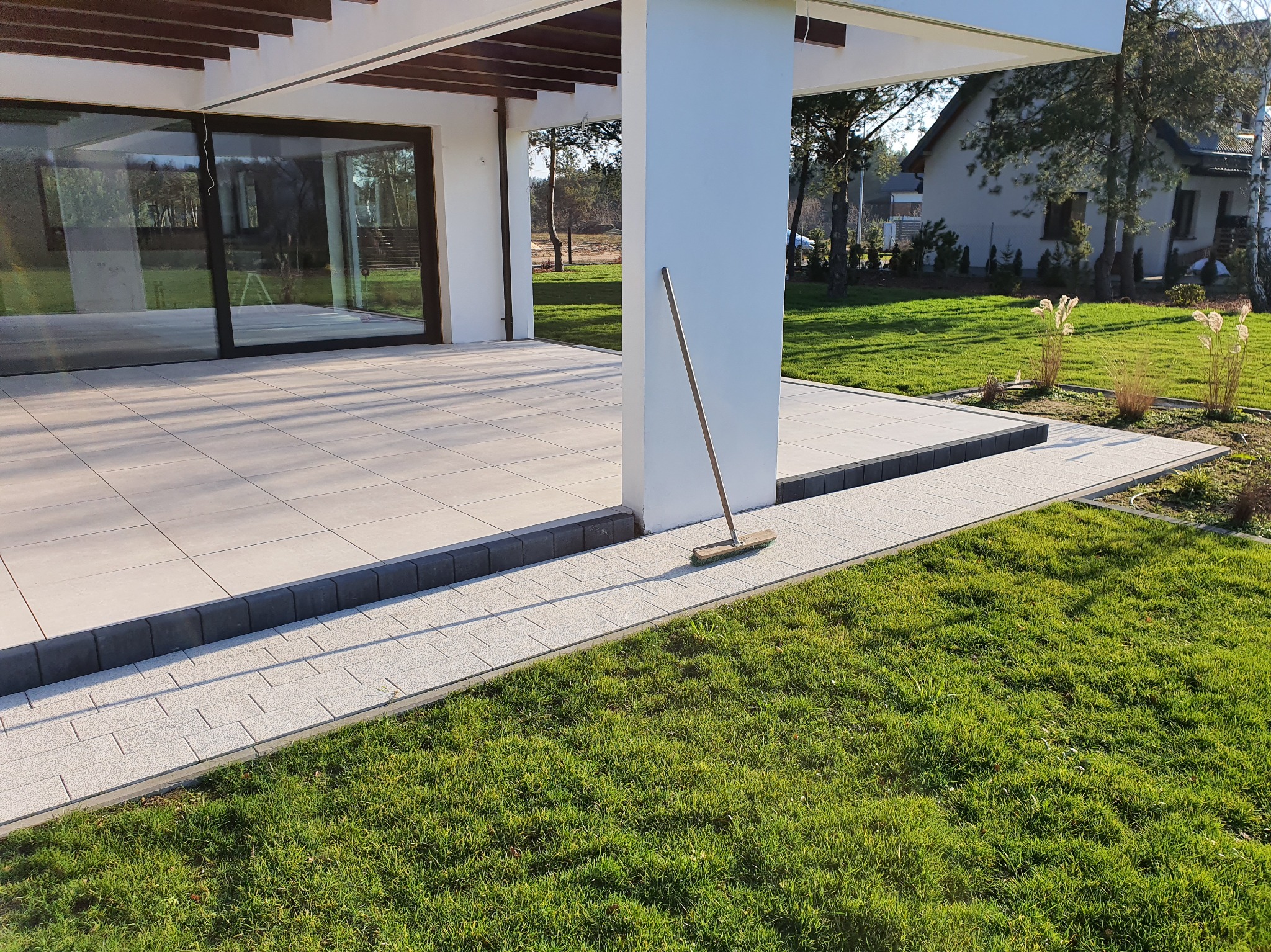 Nowoczesny taras z jasnych płyt, obrzeża z ciemnej kostki. Widoczna część domu z dużymi oknami i zadbany trawnik. Na tarasie stoi miotła.
