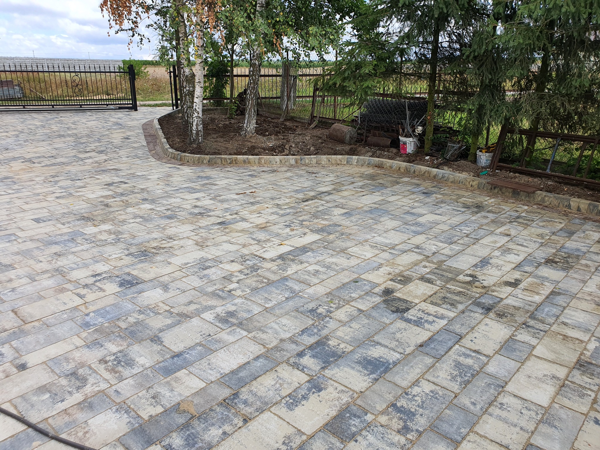 Ułożona kostka brukowa w odcieniach szarości i beżu tworzy wzór na podjeździe. Widoczne drzewa i ogrodzenie w tle. Estetyczne wykończenie obrzeży.